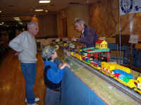 Chuck Smith (left) watches as a young visitor operates the log loader accessory.<br>George Cole (right) is reloading planks into the sawmill.